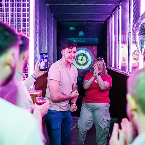 A man celebrates enthusiastically in a neon-lit axe-throwing lane with a dartboard in the background, while others clap and cheer, capturing the lively moment.