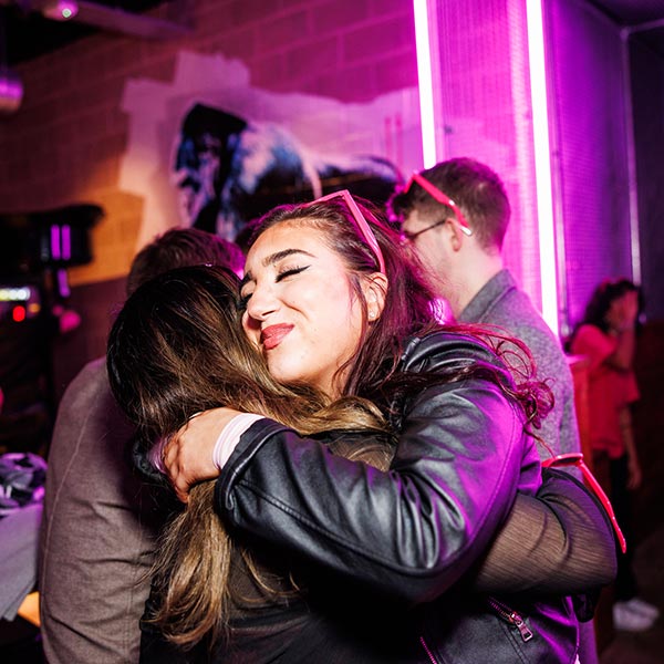 Two people embrace in a dimly lit room with neon purple lighting. Other people are in the background.