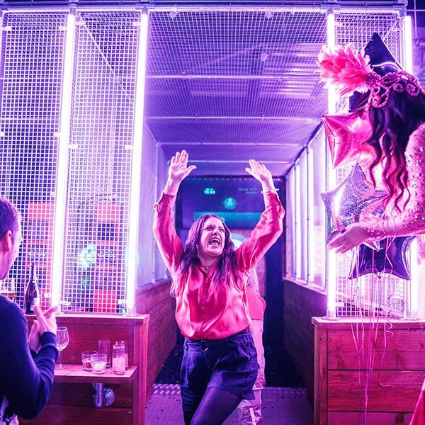 A woman dances joyfully in a neon-lit axe-throwing lane wearing a red blouse. Star-shaped balloons and drinks on a wooden ledge add to the festive atmosphere.