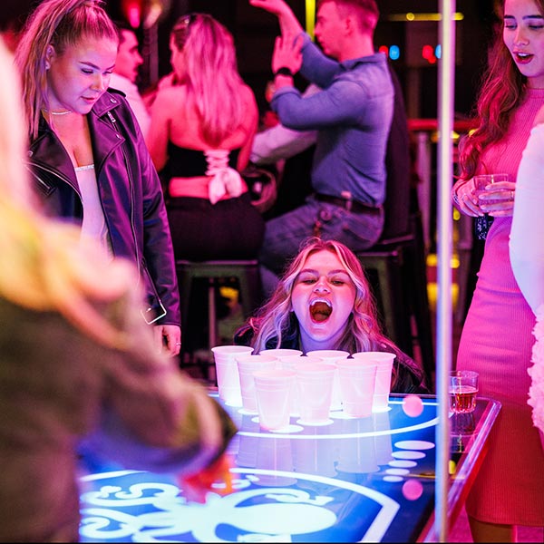 A lively party scene with a group playing a neon-lit cup game, as one woman leans forward with excitement while others watch and cheer.