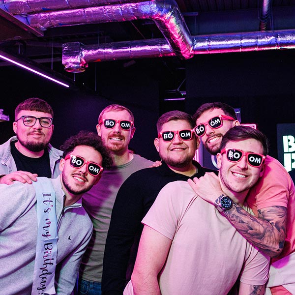 6 male party goers posing for a photo. 5 of them are wearing dress-up BOOM sunglasses. 1 is wearing a Happy Birthday sash