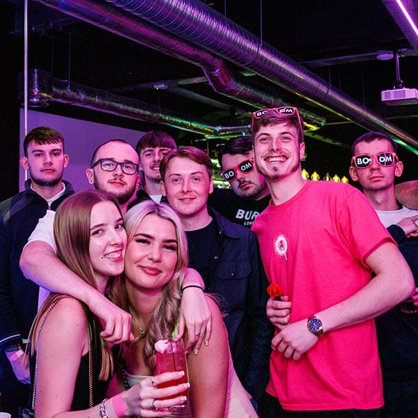 2 girls hugging in the foreground with 7 men behind them, all posing for a photo under a pink neon light.