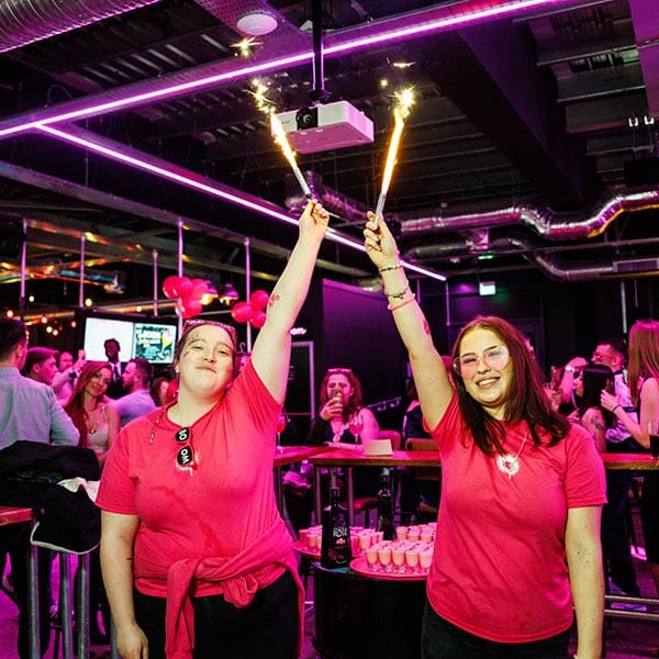 Two members of staff at BOOM Battle Bar standing with one arm in the air holding onto lit sparklers