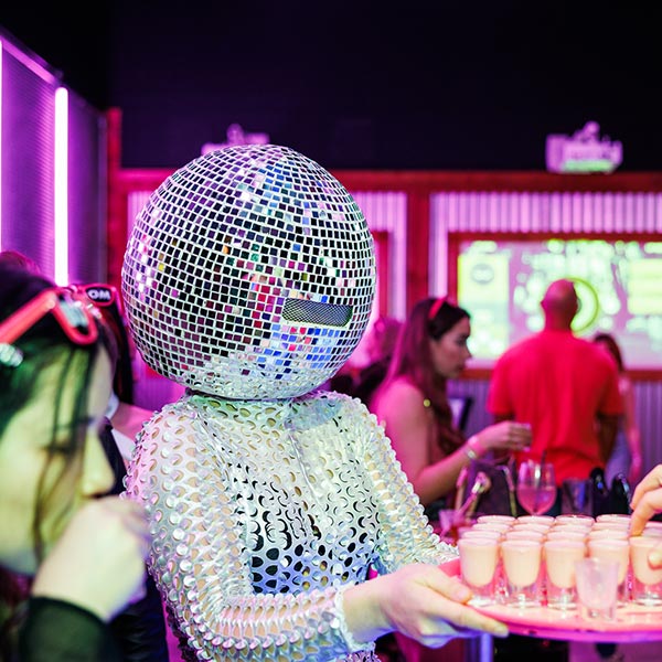 A lady wearing a large glitter ball head and sparkly costume is handing out shots on a tray