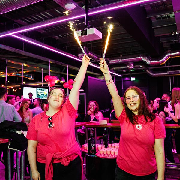 Two members of staff at BOOM Battle Bar standing with one arm in the air holding onto lit sparklers